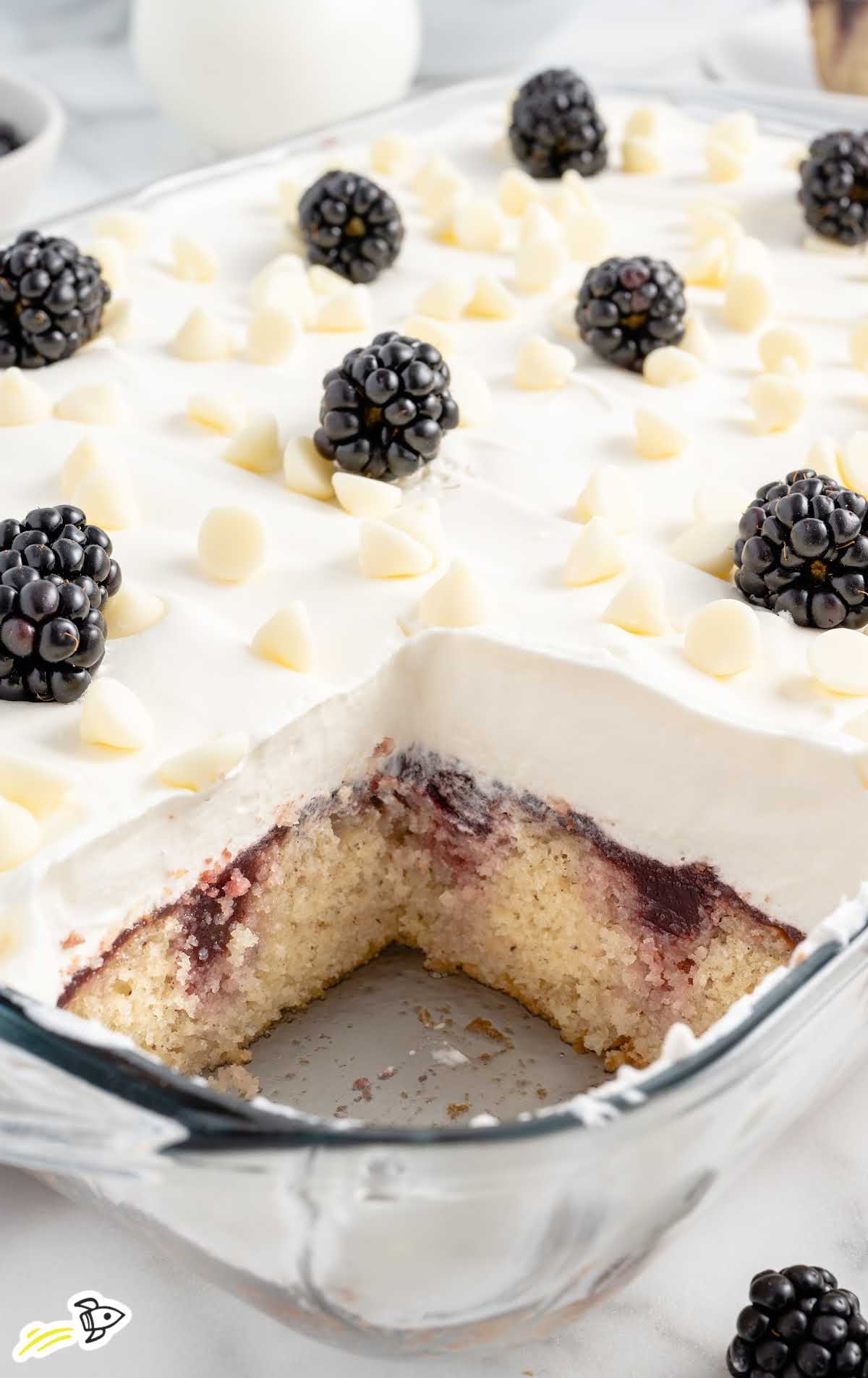 a close-up shot of White Chocolate Blackberry Poke Cake on a baking dish with a slice taken out