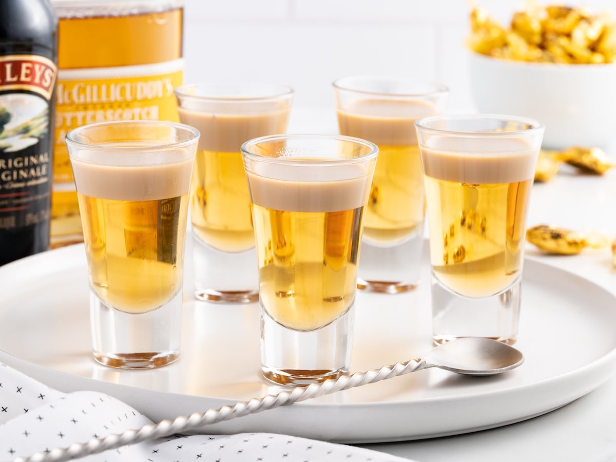 Buttery nipple shots with creamy tops are arranged on a white tray, surrounded by bottles of Baileys and liqueur. A metal spoon and a bowl of snacks add to the scene, perfect for pairing.