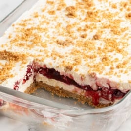 A glass dish filled with Cherry Yum Yum, a classic layered dessert featuring a crumb crust, creamy layer, cherry filling, and whipped topping, with crumbs sprinkled on top. A slice has been removed, showcasing the tempting layers of this delicious treat.