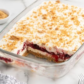 A glass dish of Cherry Yum Yum dessert with a graham cracker crust, creamy filling, cherry topping, and whipped topping. A portion is missing, revealing the delicious layers inside. Crumbs are sprinkled on top.
