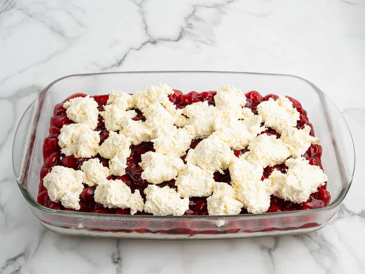 A glass baking dish filled with cherry pie filling, topped with uneven dollops of biscuit or cake batter, sits on a white marble countertop—reminiscent of a classic Cherry Yum Yum dessert.
