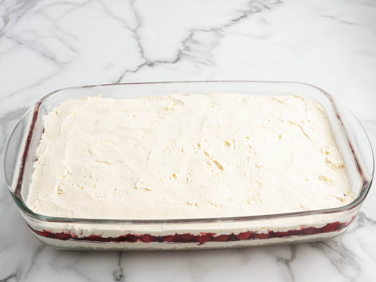 A glass baking dish filled with Cherry Yum Yum, a layered dessert featuring a creamy white top layer and a vibrant red fruit layer underneath, sits invitingly on a marble countertop.