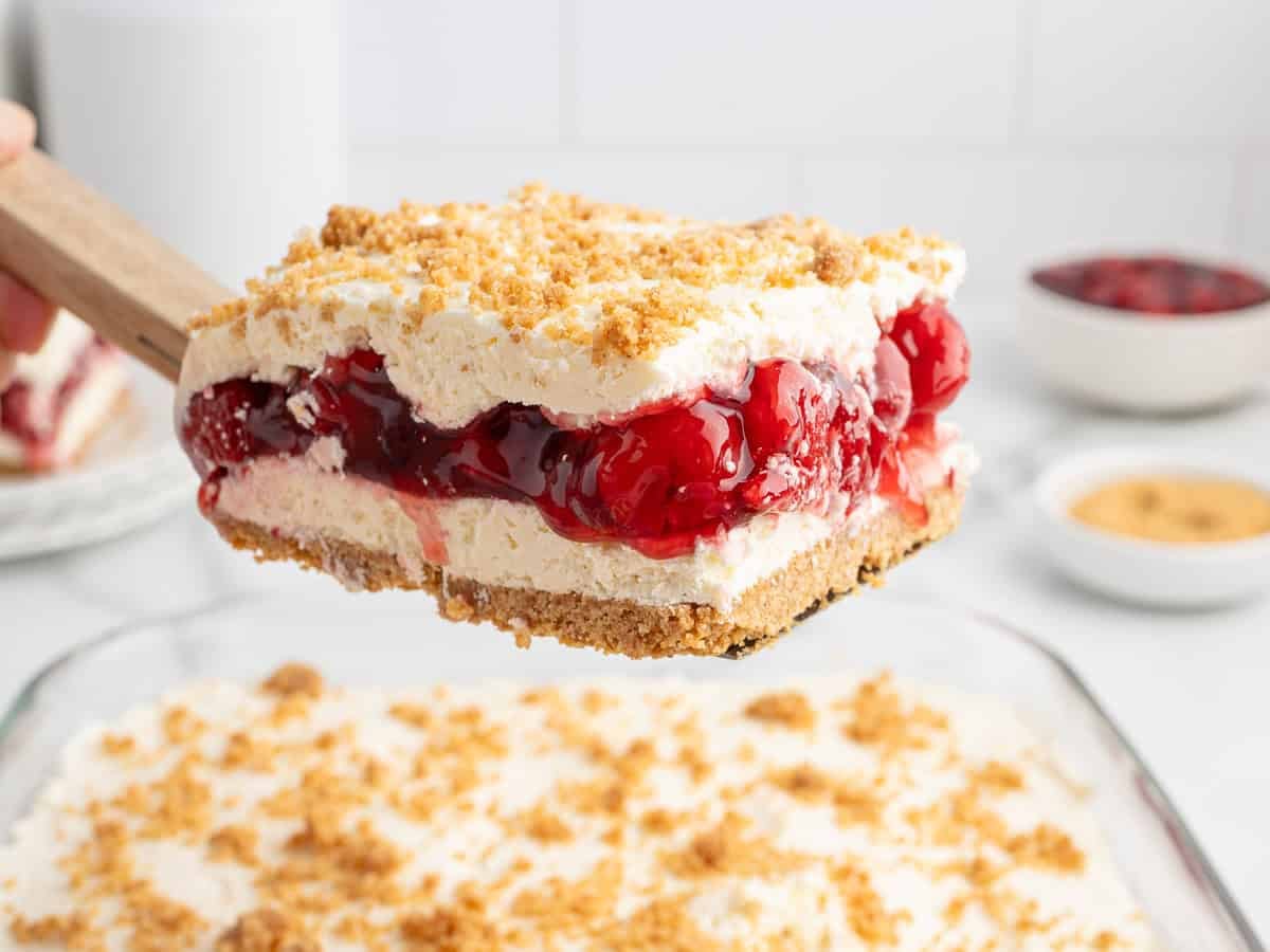 A spatula lifts a slice of Cherry Yum Yum, the layered dessert with a crumbly crust, creamy filling, cherry topping, and more crumbs, above a glass dish of the same dessert. Bowls with crumbs and cherries are in the background.