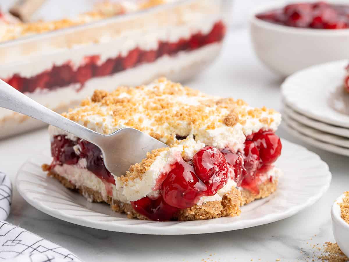 A fork lifts a slice of layered Cherry Yum Yum dessert with whipped cream and crumb topping from a white plate; the dessert has a crumb crust, creamy middle, and a sweet cherry filling. A larger dish and bowls are in the background.