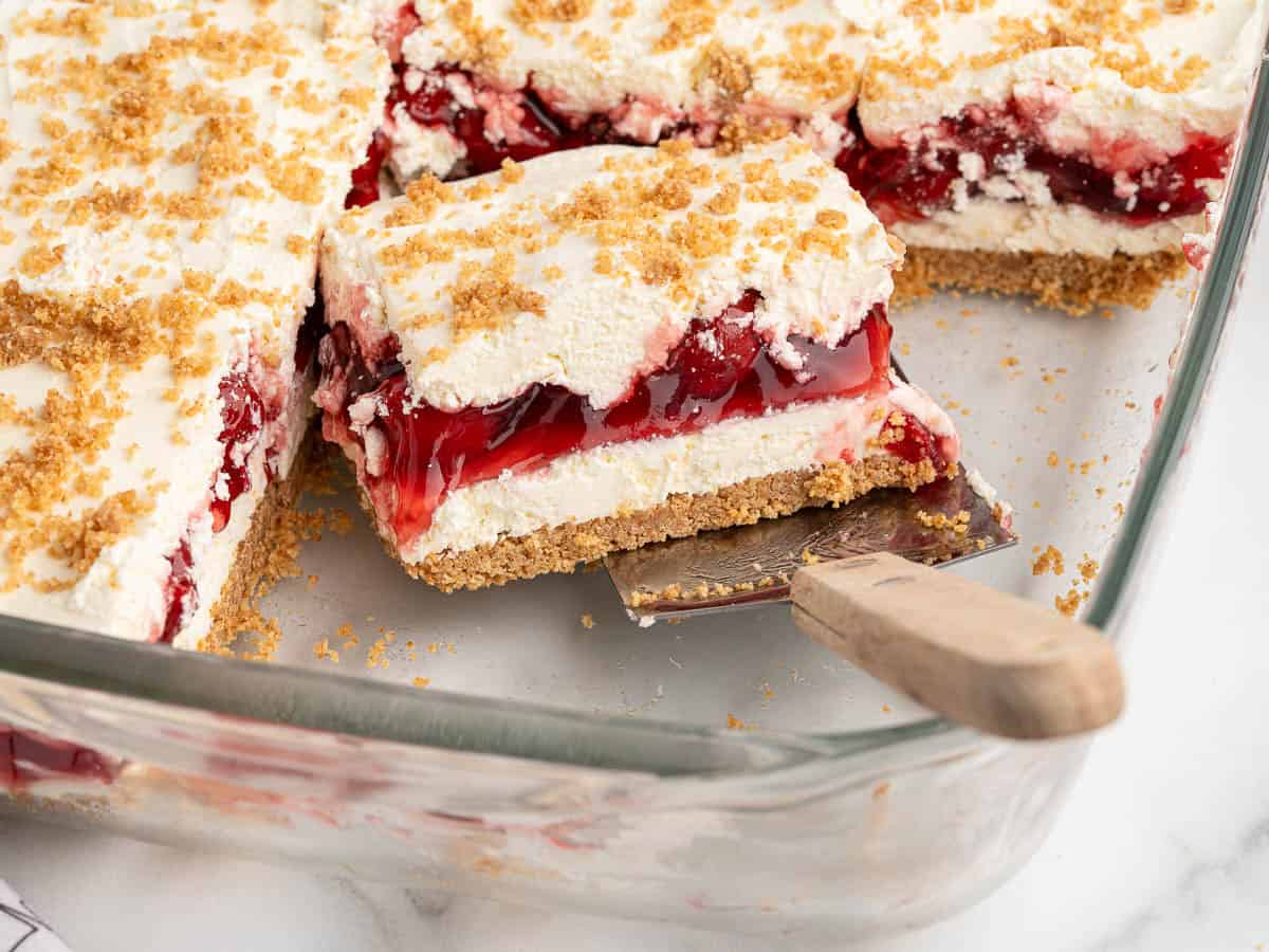 A glass dish with layered Cherry Yum Yum dessert bars featuring a crumbly crust, creamy middle layer, and cherry filling, topped with more crumbs. One bar is lifted out with a metal spatula.
