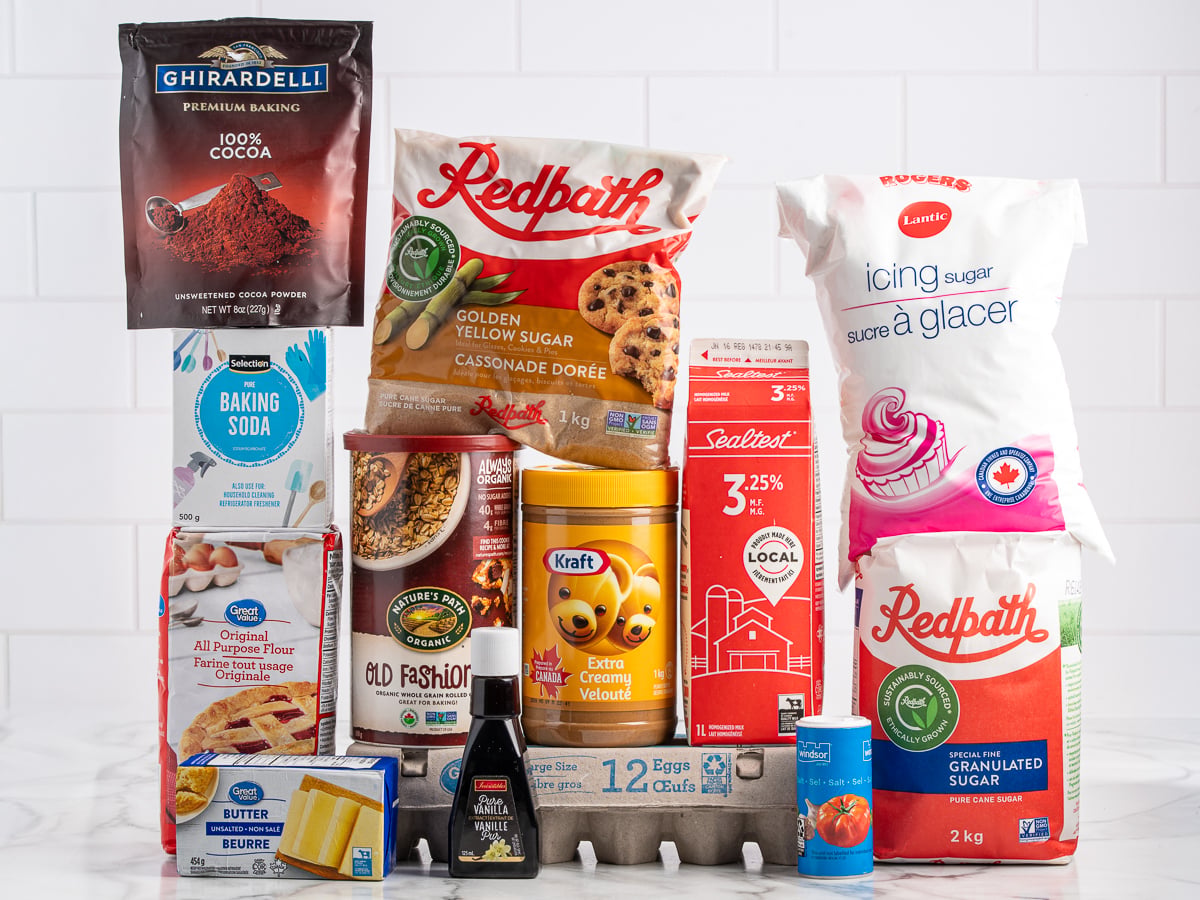 A variety of baking ingredients for Lunch Lady Peanut Butter Bars, including sugars, flour, cocoa powder, baking soda, oats, butter, eggs, vanilla extract, and milk, neatly arranged on a white countertop with a tiled background.
