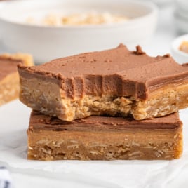 Two oatmeal bars stacked, each topped with a thick layer of chocolate frosting—just like classic Lunch Lady Peanut Butter Bars. The top bar has a large bite taken out, revealing a soft, chewy texture. Blurred bowls and more bars fill the background.