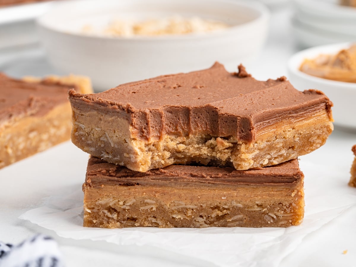 Two oatmeal bars stacked, each topped with a thick layer of chocolate frostingโjust like classic Lunch Lady Peanut Butter Bars. The top bar has a large bite taken out, revealing a soft, chewy texture. Blurred bowls and more bars fill the background.