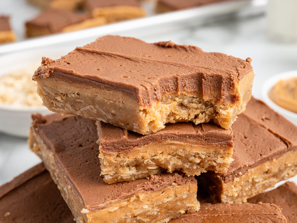 Stack of Lunch Lady Peanut Butter Bars with a thick chocolate and peanut butter topping, one with a bite taken out. Other bars and bowls of ingredients are visible in the background.