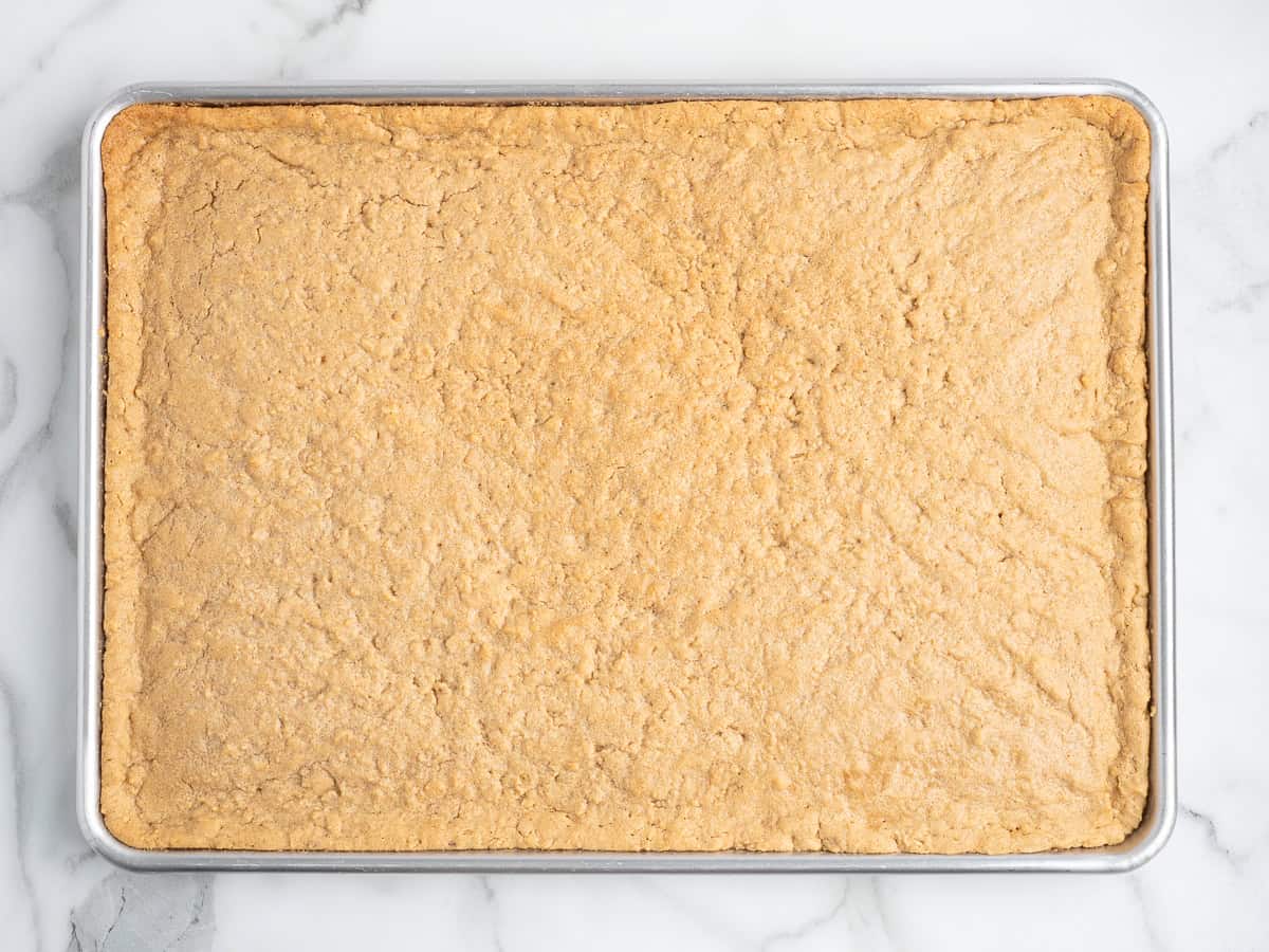 A rectangular pan filled with a large, uncut sheet of golden-brown Lunch Lady Peanut Butter Bars rests on a white marble surface.