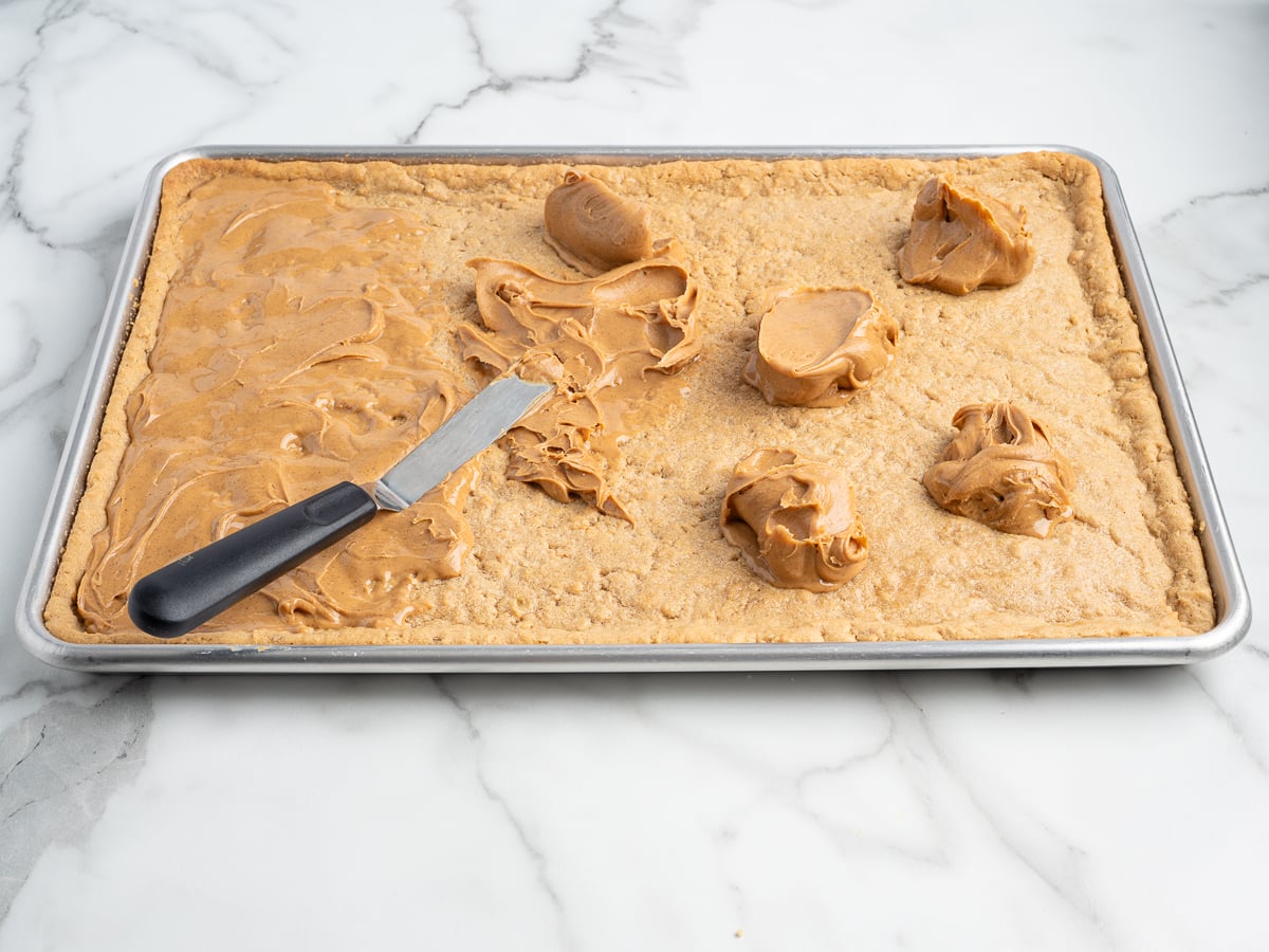 A baking sheet with a large rectangular layer of cookie dough for Lunch Lady Peanut Butter Bars, topped with several scoops of peanut butter, partially spread with an offset spatula on a marble countertop.
