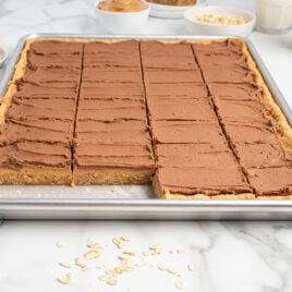 A tray of frosted Lunch Lady Peanut Butter Bars cut into squares, with one square missing. Ingredients like oats and peanut butter, as well as glasses of milk, are visible in the background on a marble surface.