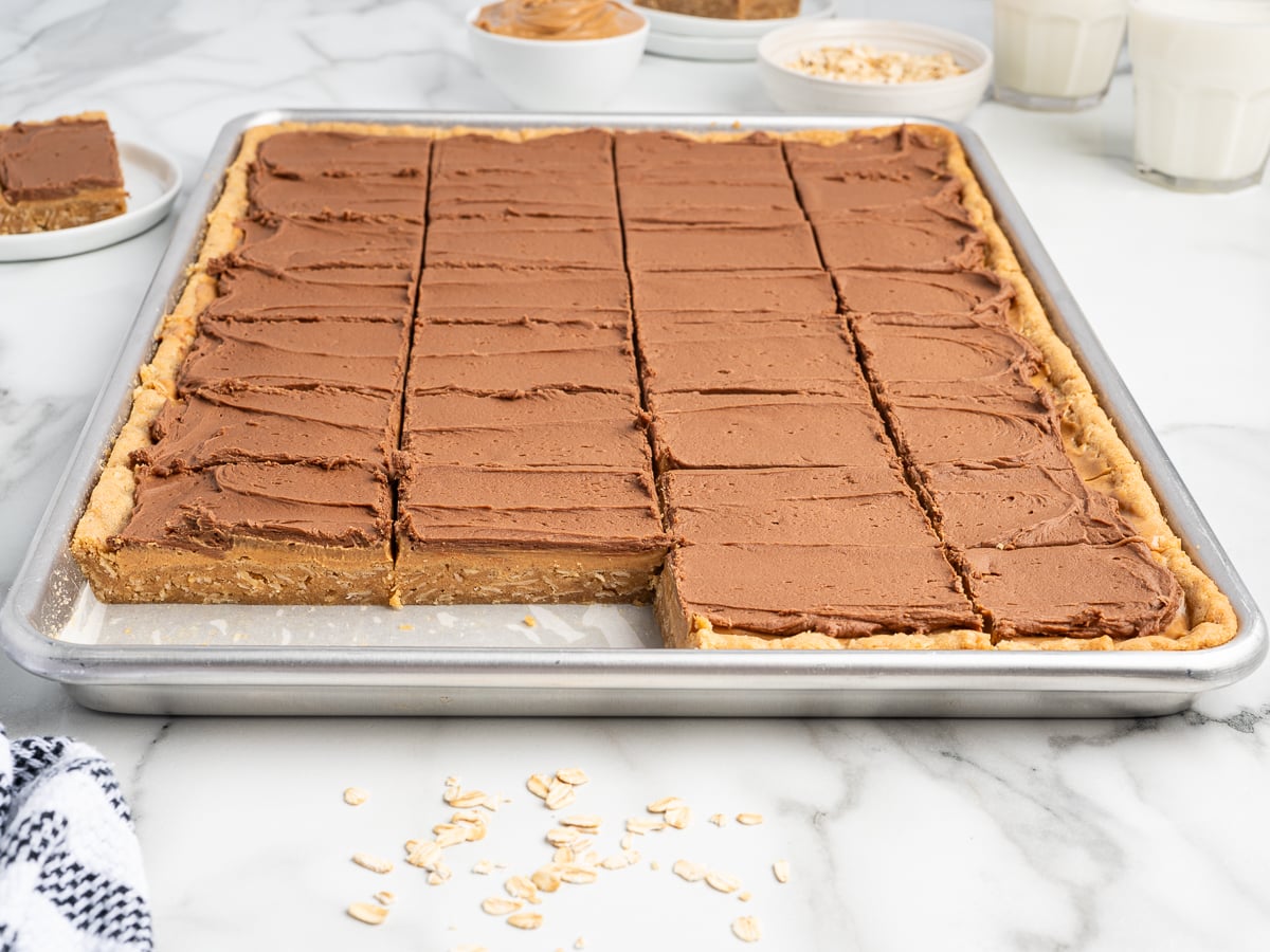 A tray of frosted Lunch Lady Peanut Butter Bars cut into squares, with one square missing. Ingredients like oats and peanut butter, as well as glasses of milk, are visible in the background on a marble surface.