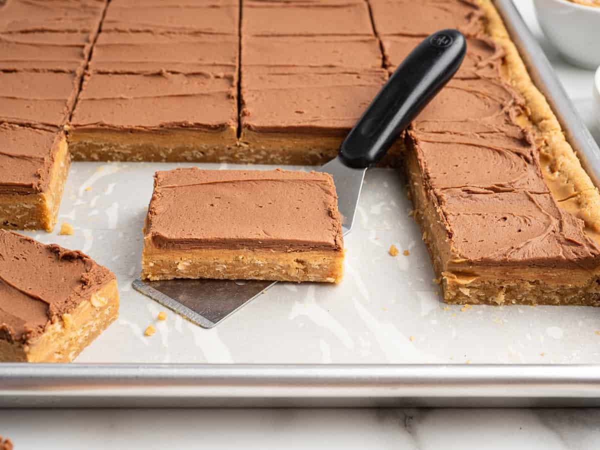 A tray of Lunch Lady Peanut Butter Bars cut into squares, with one bar lifted by a spatula. These bars feature a light, dense base and are topped with a smooth layer of chocolate frosting.
