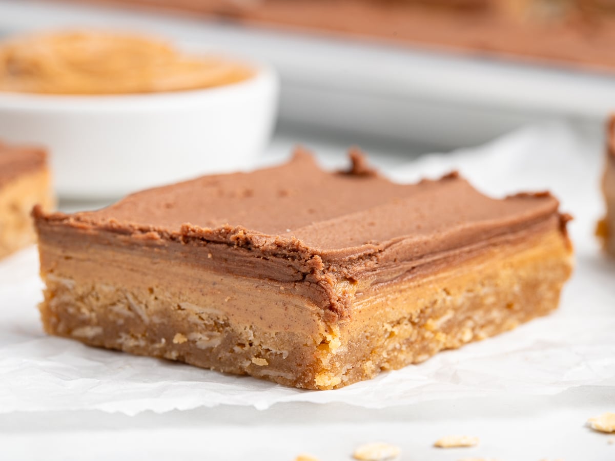A close-up of Lunch Lady Peanut Butter Bars with three distinct layers: a crumbly oat base, a creamy peanut butter middle, and a smooth chocolate top, resting on white parchment paper.