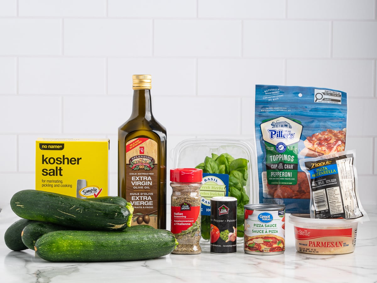 Several ingredients for Pizza Zucchini Boats are arranged on a marble counter, including zucchinis, kosher salt, extra virgin olive oil, dried Italian seasoning, fresh basil, pizza sauce, pepperoni slices, shredded mozzarella, and grated parmesan cheese.