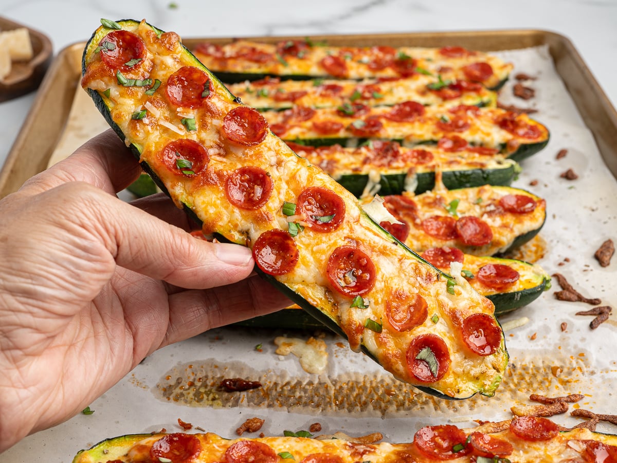 A hand holds a Pizza Zucchini Boat topped with melted cheese and mini pepperoni slices, with more Pizza Zucchini Boats on a parchment-lined baking sheet in the background.