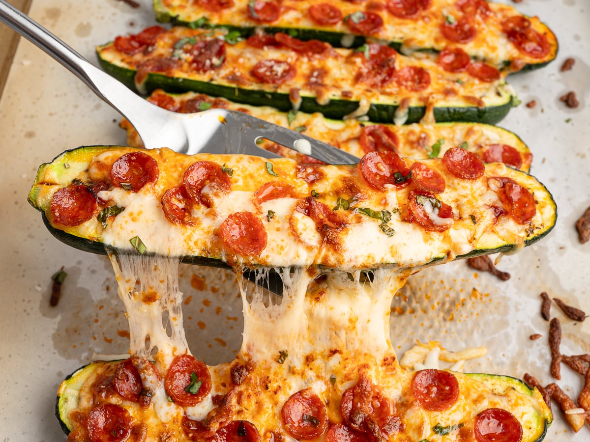 A close-up of Pizza Zucchini Boats topped with melted cheese and mini pepperoni slices, being lifted from a baking sheet with a spatula, showcasing stretchy, gooey cheese.