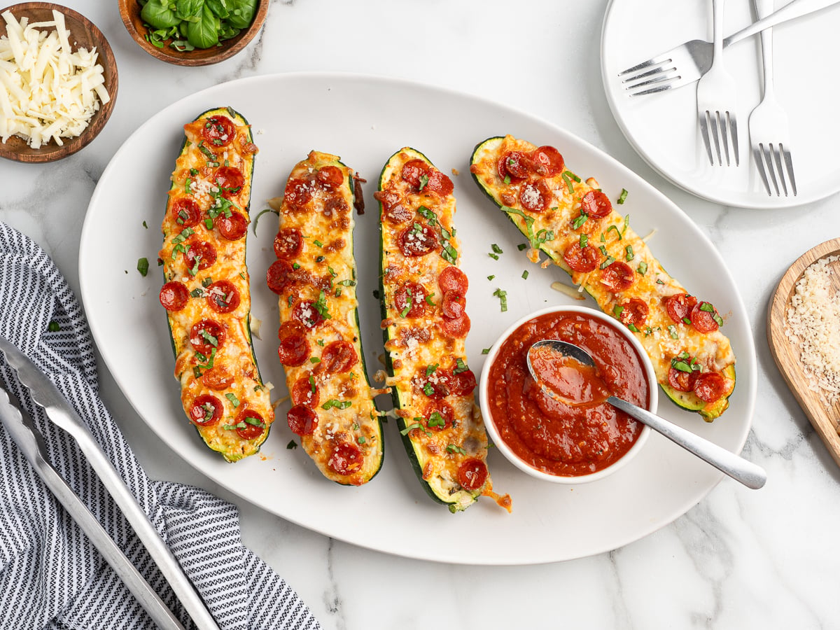 A white plate with four Pizza Zucchini Boats topped with melted cheese and mini pepperoni slices, served with a bowl of marinara sauce. Plates, forks, and bowls of cheese and basil rest nearby on a marble surface.