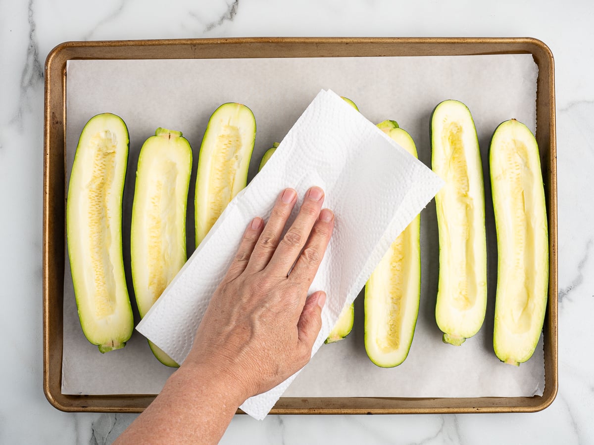 A hand uses a paper towel to pat dry halved zucchinis for Pizza Zucchini Boats, placed cut side up on a parchment-lined baking sheet.