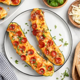 Two Pizza Zucchini Boats on a plate, topped with melted cheese, mini pepperoni slices, and chopped herbs. Surrounding the plate are bowls of shredded cheese, grated parmesan, fresh basil, and a striped napkin.
