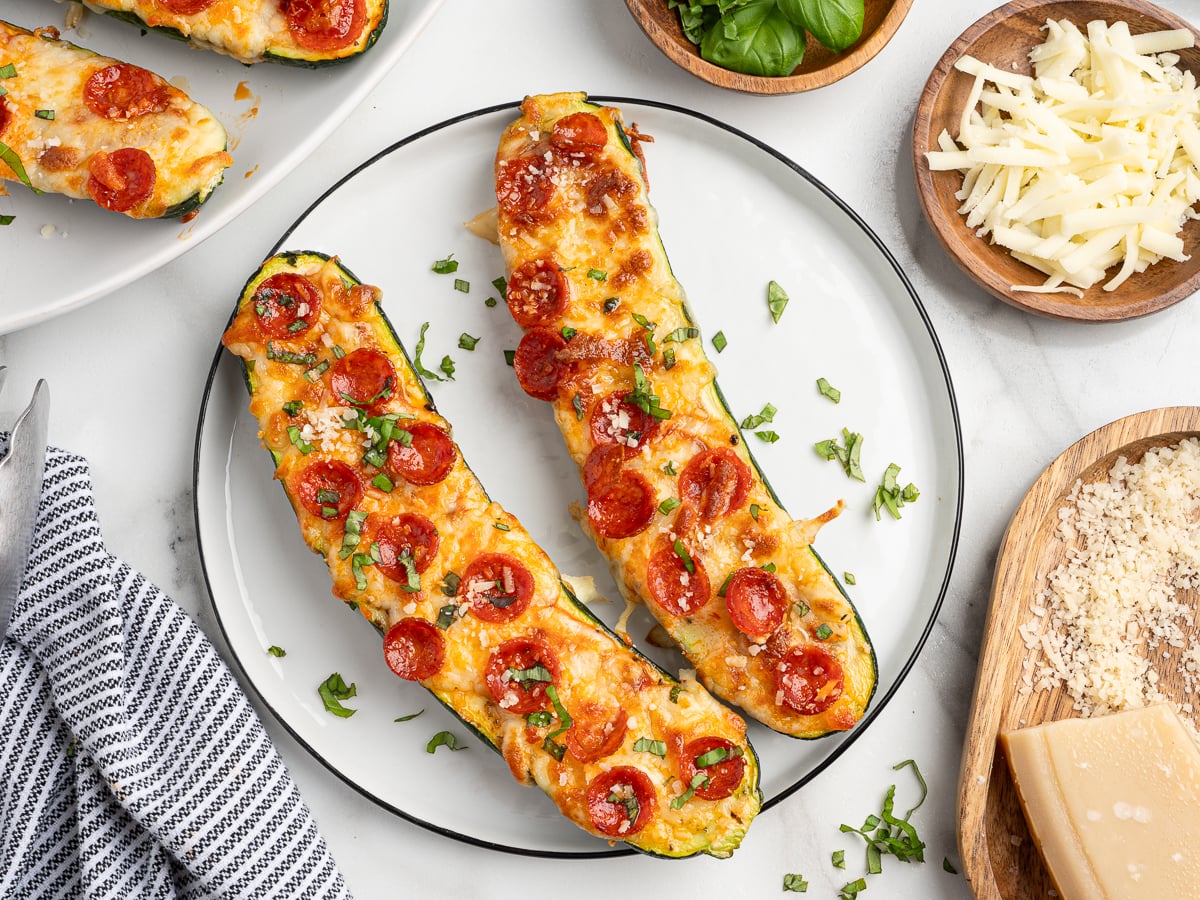 Two Pizza Zucchini Boats on a plate, topped with melted cheese, mini pepperoni slices, and chopped herbs. Surrounding the plate are bowls of shredded cheese, grated parmesan, fresh basil, and a striped napkin.