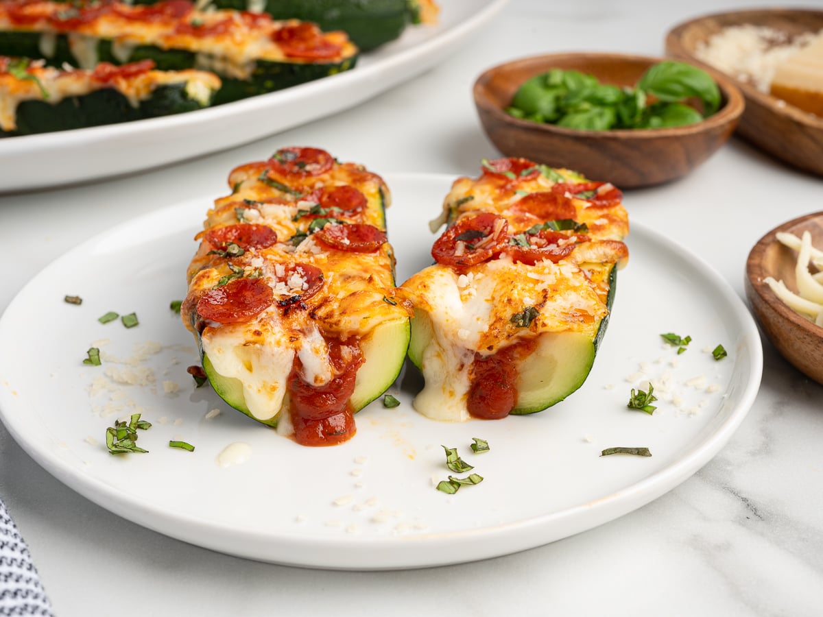 Two Pizza Zucchini Boats rest on a white plate, filled with melted cheese, tomato sauce, mini pepperoni slices, and chopped herbs. Small bowls of grated cheese and fresh herbs sit in the background.