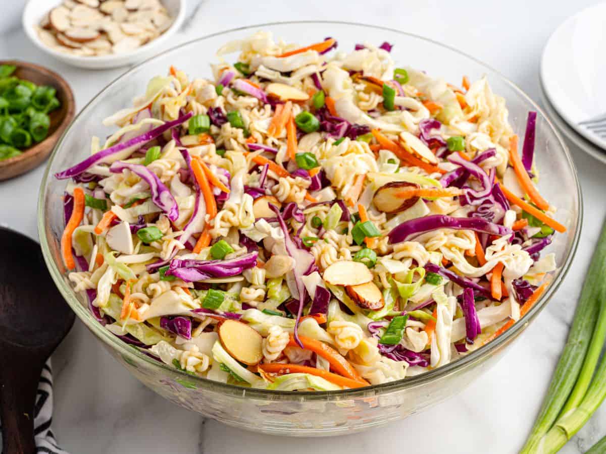 A glass bowl filled with colorful Ramen Noodle Salad sits on a marble surface, showcasing shredded cabbage, carrots, purple cabbage, green onions, sliced almonds, and crunchy ramen noodles. Small plates of toppings are nearby to complete this vibrant Asian slaw.