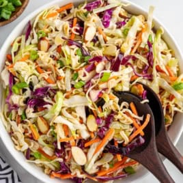A bowl of colorful Ramen Noodle Salad with shredded carrots, sliced almonds, green onions, and crunchy ramen noodles, served with two dark wooden salad utensils.