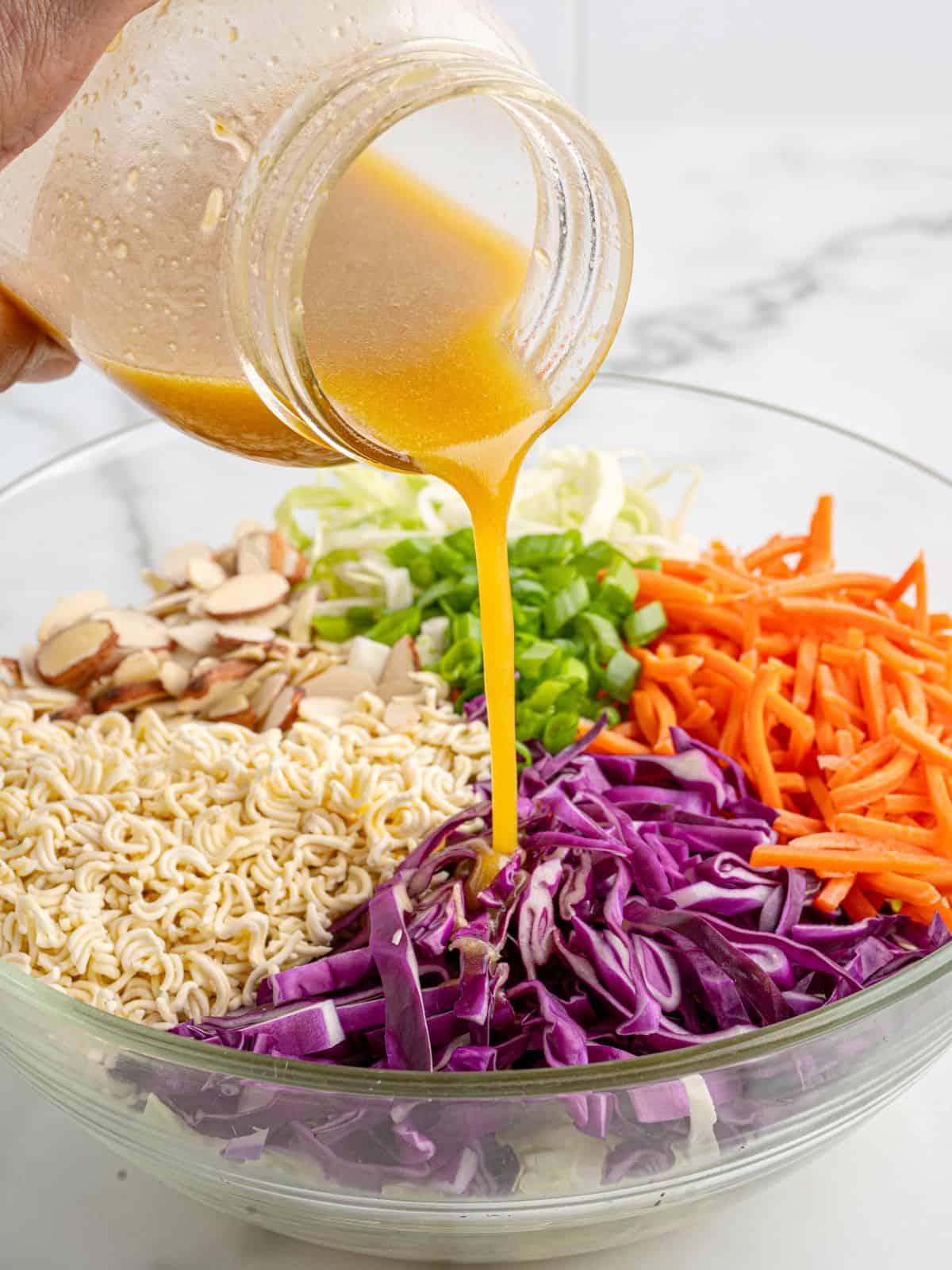 A hand pours orange dressing from a jar onto a bowl of Ramen Noodle Salad containing shredded cabbage, carrots, green onions, sliced almonds, and uncooked ramen noodles.