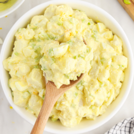 A white serving bowl of Amish Potato Salad, with a wooden spoon lifting out a scoop.