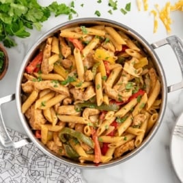 Overhead view of a stainless steel pot filled with chicken fajita pasta made with penne, sliced bell peppers, and strips of seasoned chicken, garnished with chopped cilantro. Surrounding the pot are a bowl of shredded cheese, a bowl of chopped herbs, a stack of white plates with forks, and fresh cilantro on a white marble surface.