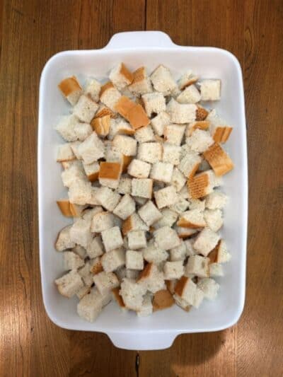 White baking dish filled with cubed pieces of white bread, some with crusts, sitting on a wooden tabletop—perfect for making a delicious Pumpkin French Toast Casserole.