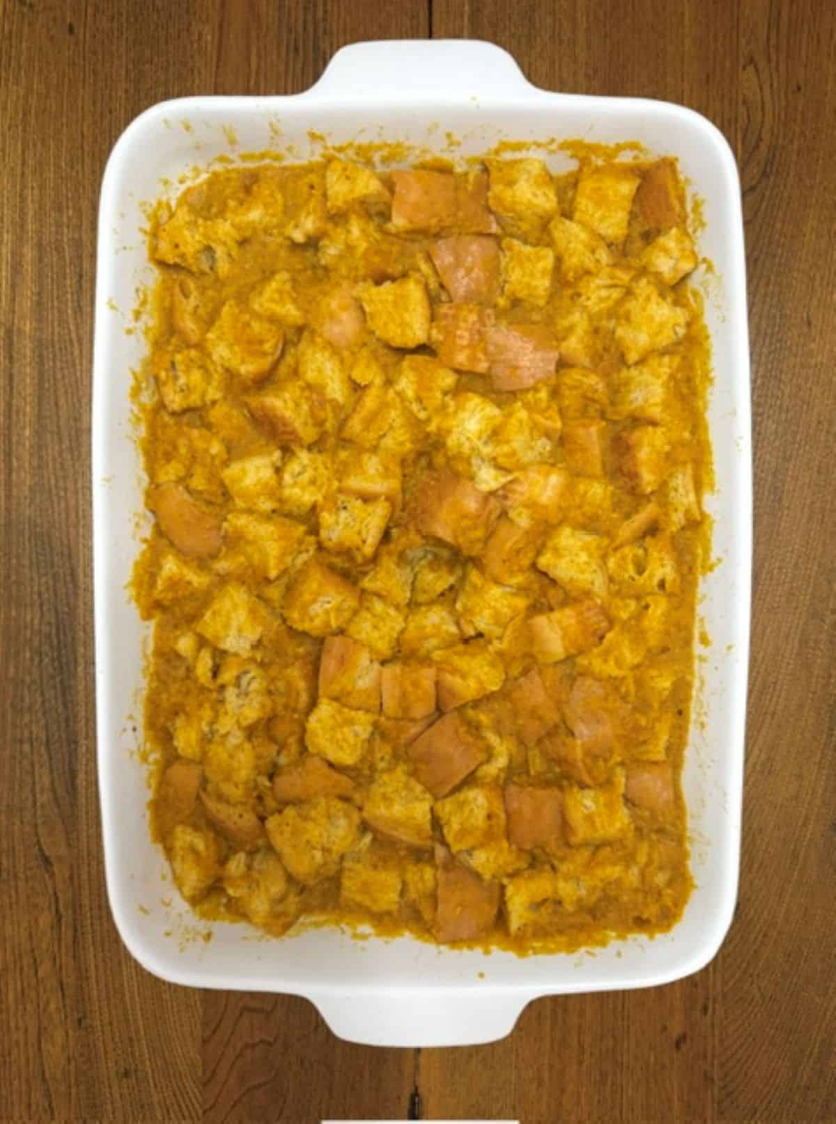 A white baking dish filled with cubed bread mixed with a pumpkin-orange mixture, sitting on a wooden surface, ready to be baked into a delicious Pumpkin French Toast Casserole.