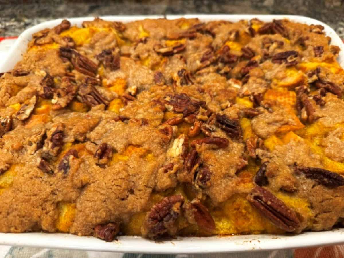 A Pumpkin French Toast Casserole topped with a crumbly brown sugar and pecan streusel sits in a white rectangular dish on the countertop.