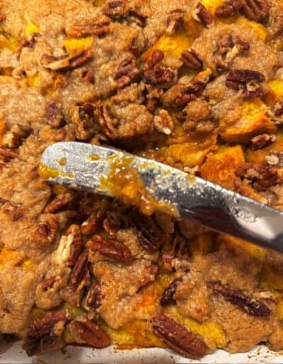 A close-up of a pecan-topped coffee cake, with a butter knife spreading a gooey portion. Reminiscent of the cozy flavors in Pumpkin French Toast Casserole, the topping looks crumbly with visible pecans, and the cake appears moist and golden underneath.
