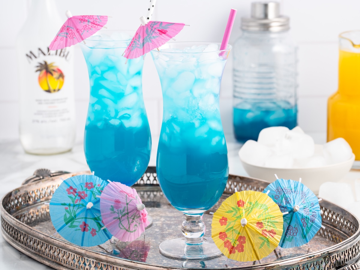 Blue ocean drink recipe in hurricane glasses with pink cocktail umbrellas and a pink straw on a silver tray