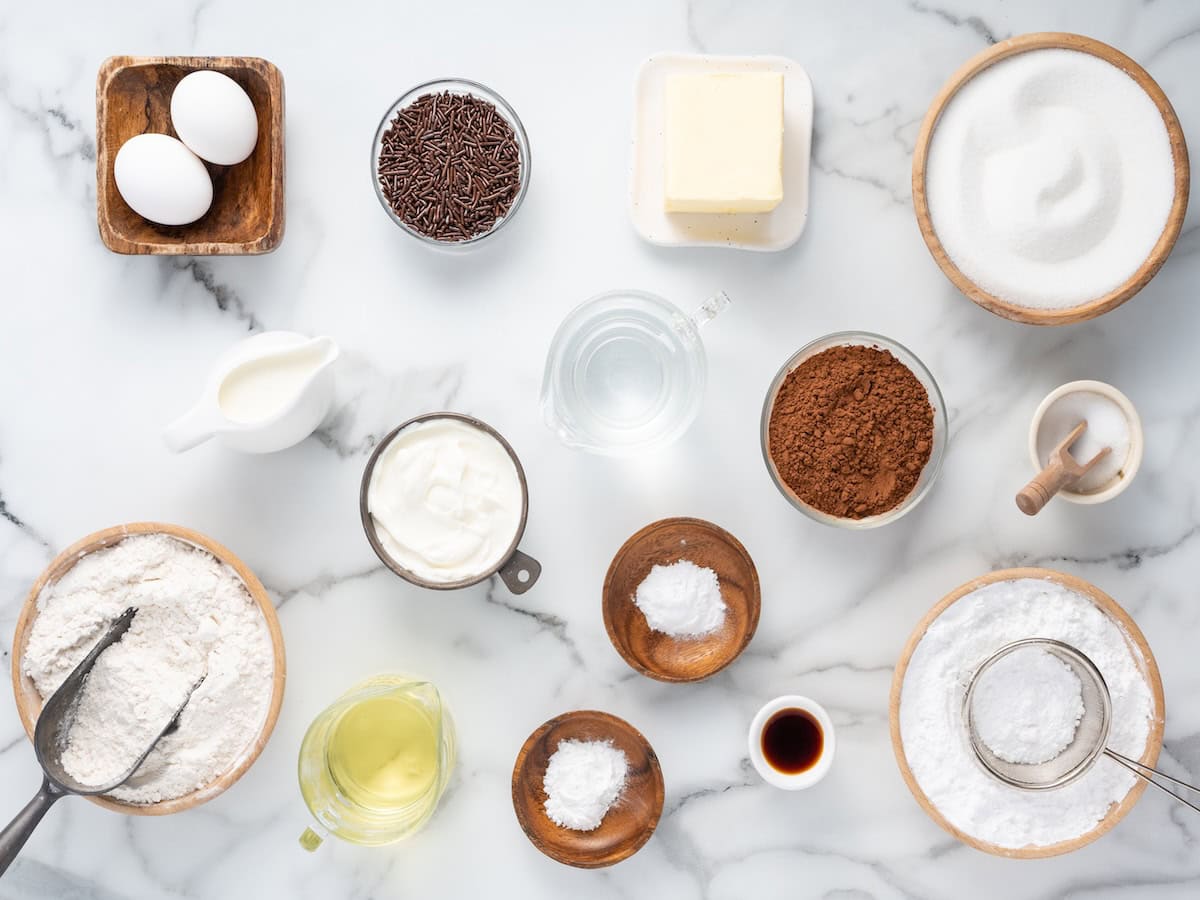 Baking ingredients on a marble surface: eggs, chocolate sprinkles, butter, sugar, milk, flour, oil, cocoa powder, vanilla, baking powder, baking soda, and salt—all carefully arranged in bowls and cups—perfectly set the scene for crafting a moist chocolate cupcake recipe.