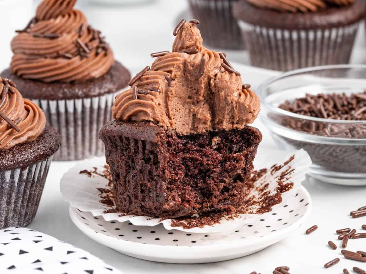 A moist chocolate cupcake—crafted from a beloved Moist Chocolate Cupcake Recipe—with rich chocolate frosting and sprinkles sits on a plate, partially unwrapped and missing a large bite. Other cupcakes and a bowl of chocolate sprinkles are in the background.
