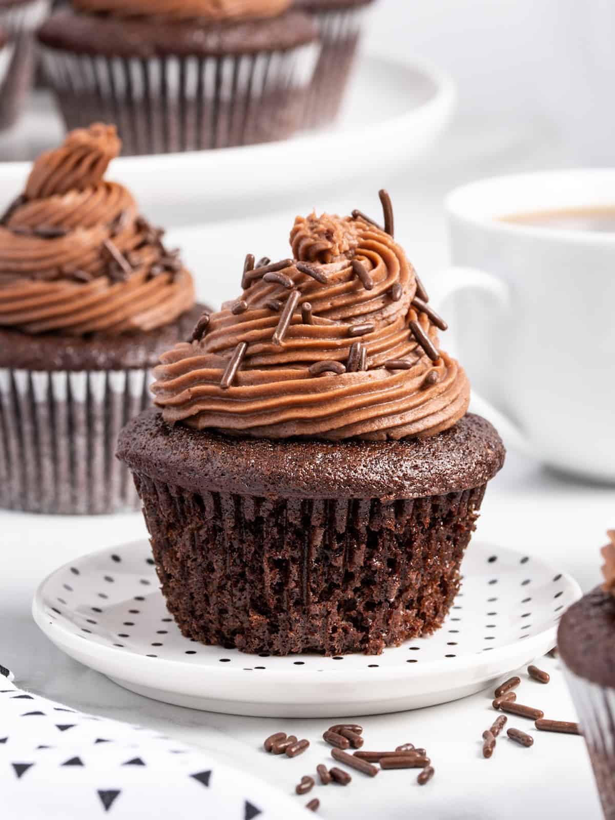Moist chocolate cupcake with rich chocolate frosting and colorful sprinkles on a white plate. Other cupcakes and a cup of coffee are blurred in the background, showcasing this delightful treat made from a signature moist chocolate cupcake recipe.