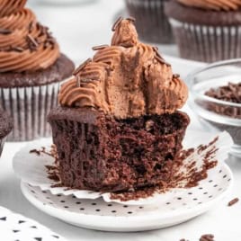 A chocolate cupcake with rich chocolate frosting and colorful sprinkles sits on a plate with a bite taken out, revealing its perfectly moist inside—a delicious example of the perfect Moist Chocolate Cupcake Recipe. In the background, other cupcakes and a bowl of chocolate sprinkles complete the tempting scene.