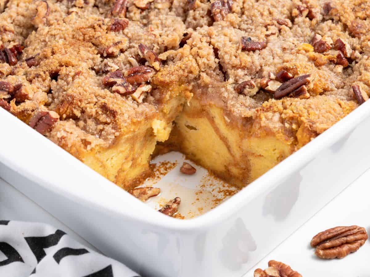 Pumpkin French Toast Casserole baked to perfection, topped with pecans and crumbly streusel, is shown in a white dish with one piece removed, revealing the soft, golden inside.