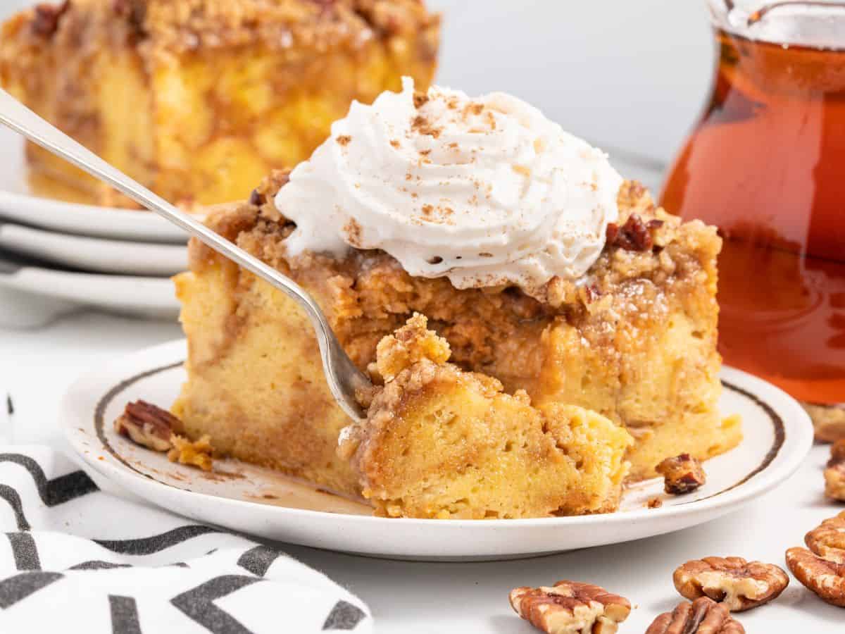A fork cuts into a slice of Pumpkin French Toast Casserole topped with whipped cream on a white plate, surrounded by pecans and a syrup pitcher.