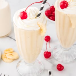 Two tall glasses of creamy banana milkshake topped with whipped cream, maraschino cherries, and black-and-white striped straws, garnished with banana slices, with extra cherries and banana chips on a white marble surface.