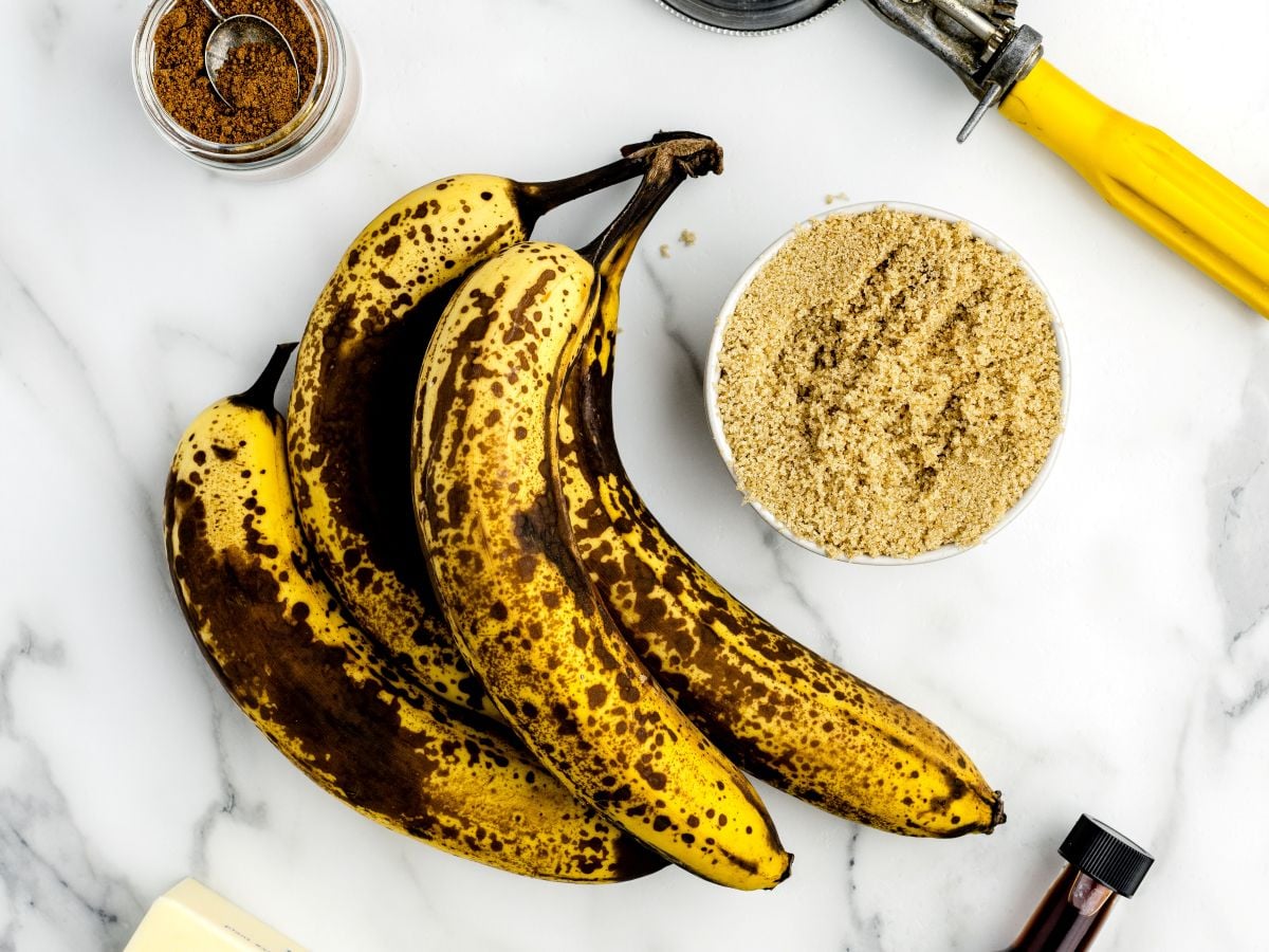 Three ripe bananas with brown spots rest on a white surface next to a bowl of brown sugar, ready to become caramelized bananas with the help of cinnamon, vanilla extract, and a yellow-handled tool.