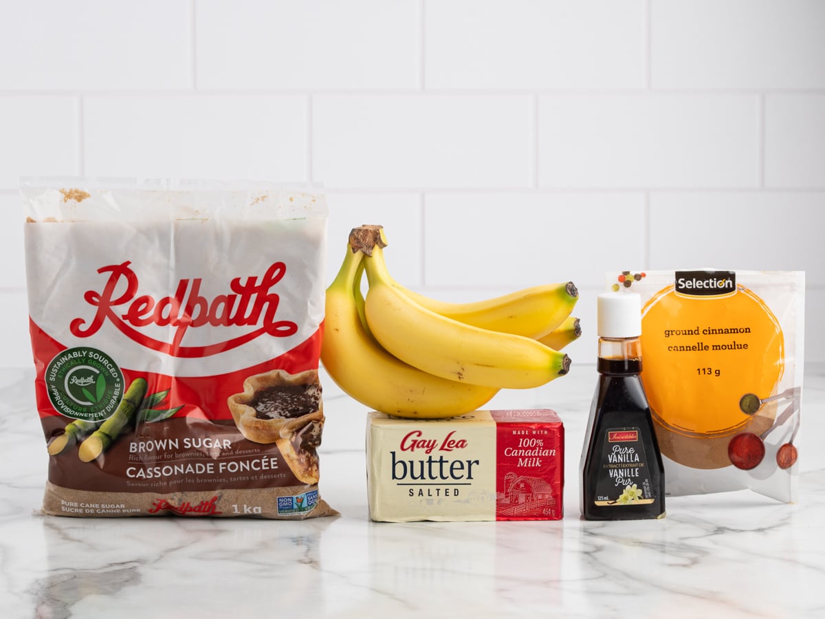 A bag of Redpath brown sugar, a bunch of bananas, a block of salted butter, vanilla extract, and ground cinnamon&mdash;everything you need for delicious caramelized bananas&mdash;set on a white marble counter with a white tile background.