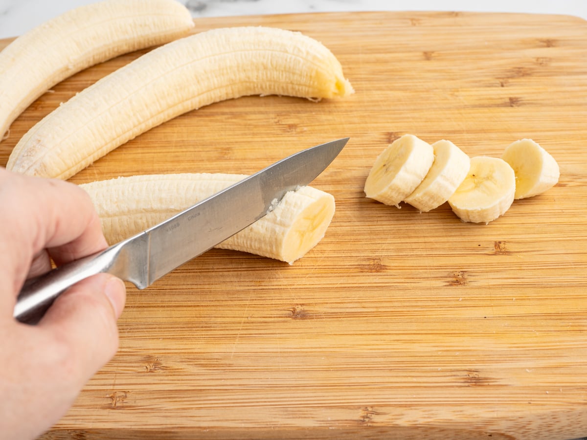 A hand uses a knife to slice a peeled banana on a wooden cutting board, preparing caramelized bananas. Several banana slices and two uncut bananas are also visible on the board.