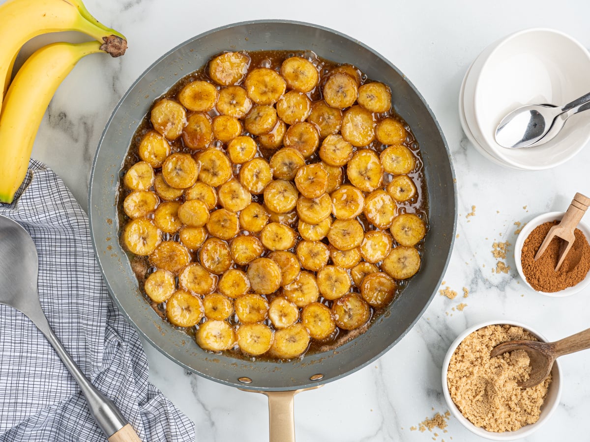 A skillet filled with Caramelized Bananas in sauce sits on a marble countertop, surrounded by fresh bananas, a gray kitchen towel, a bowl of brown sugar, cinnamon, and empty white bowls with spoons.