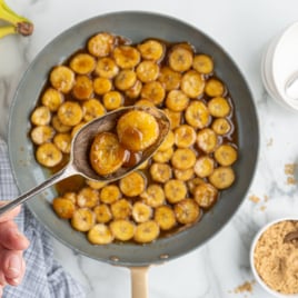 A hand holds a spoonful of caramelized bananas above a pan brimming with more golden caramelized bananas. Fresh bananas, brown sugar, cinnamon, white bowls, and a checkered cloth surround this tempting dessert scene.