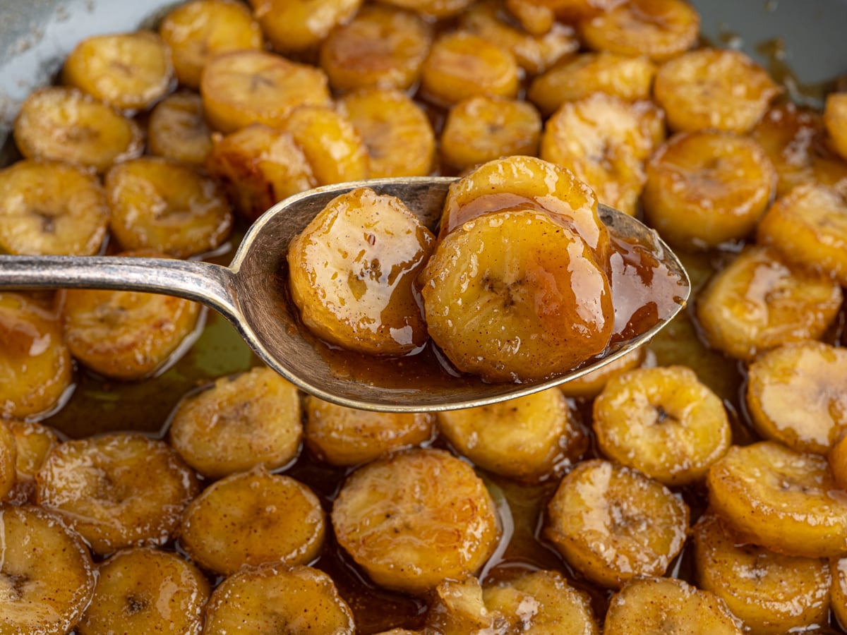 A close-up of Caramelized Bananas on a spoon, held above a pan filled with more banana slices glistening in a rich, glossy caramel sauce.